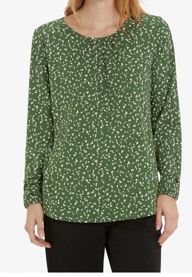 Femme portant un chemisier vert à manches longues avec un petit motif floral blanc et un pantalon noir, debout devant un fond blanc.