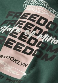T-shirt verte arborant un graphique central avec "LIBERTÉ" en noir et rose, un texte brodé "état d'esprit", et "BROOKLYN" en bas.