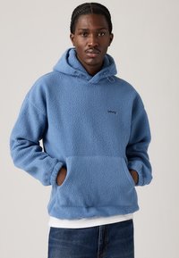 Hoodie en polaire bleue avec une poche kangourou, capuche réglable avec cordon de serrage et logo brodé. Texture douce et coupe ample.