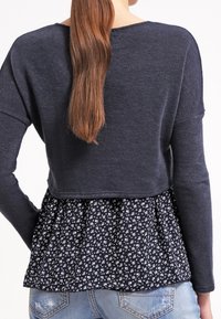 Marinblå kortad tröja med långa ärmar över en svart blommig peplum. Mjuk textur med en åtsittande överdel och en utfläkt nedre del.