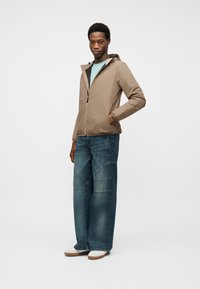 Νέος άνδρας που στέκεται με το ένα χέρι στην τσέπη, φοράει μπεζ μπουφάν με κουκούλα, ανοιχτό μπλε πουκάμισο, τζιν παντελόνι με φαρδιά μπατζάκια και λευκά αθλητικά παπούτσια.