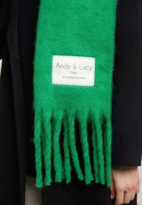 Écharpe en laine verte avec des bords à franges, portant une étiquette blanche avec "Andy & Lucy" et "Enveloppé d'Amour" en texte noir.