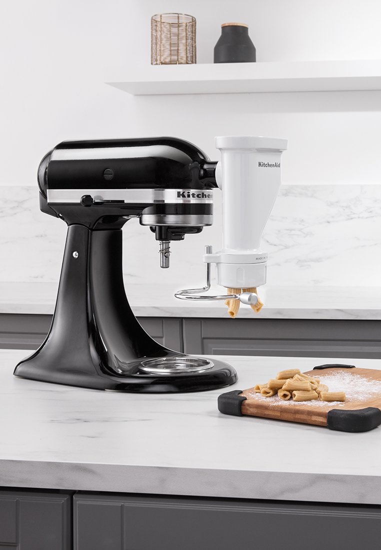 Mikser planetarny KitchenAid w czarnym metalu o błyszczącej wykończeniu, podłączony do białej ekstrudera do makaronu, ustawiony na marmurowym blacie z ciastem na drewnianej desce.