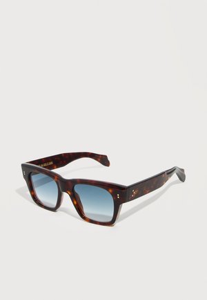 Τετράγωνα γυαλιά ηλίου με χελώνα και παχιά πλαίσια, με μπλε φακούς με διαβάθμιση, που ακουμπούν σε λευκή επιφάνεια.