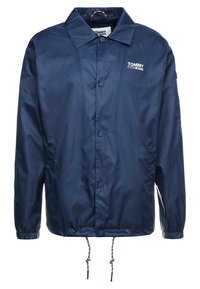 Chaqueta cortavientos azul marino hecha de un material liso y impermeable. Cuenta con un cuello con solapa, botones en la parte delantera y un dobladillo ajustable con cordón.