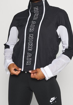 Personne qui ferme une veste coupe-vent Nike Air noire et blanche, portant des leggings Nike Air noirs avec des logos blancs.