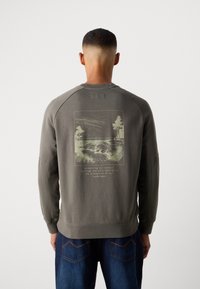 Homme avec les cheveux courts portant un sweat-shirt gris avec un imprimé de paysage et un message de conservation, associé à un jean bleu, tourné dans l'autre sens.