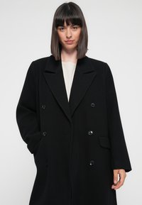 Cappotto doppiopetto nero realizzato in misto lana. Presenta ampi revers, tasche laterali e dettagli con bottoni. Tessuto morbido e vestibilità rilassata.