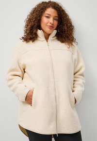 Giacca in pile beige chiaro con colletto alto, zip frontale, maniche lunghe, tasche laterali e orlo leggermente curvo. Texture morbida in tutto.