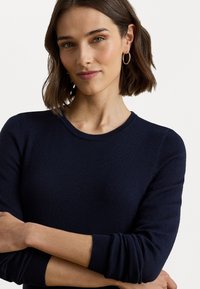 Mørkeblå strikkegenser med rund hals og lange ermer, har en glatt tekstur og en tettsittende design. Diskrete ribbede detaljer ved mansjettene.