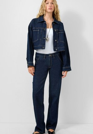 Jonge vrouw die een donkerblauw cropped spijkerjasje draagt, bijpassende hoge taille spijkerbroek, witte top en een hartvormige hangerketting.