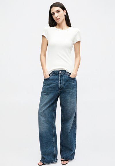 Femeie cu păr lung și închis la culoare, purtând un tricou alb cu mâneci scurte mulat, blugi albaștri cu picioare largi și sandale negre cu degetele descoperite, stând cu mâinile în buzunare.