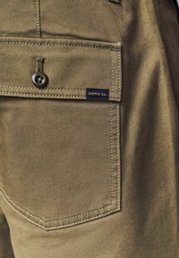 Pantaloni in cotone verde oliva con una tasca a toppa dotata di bottone e un'etichetta in tessuto blu navy con la scritta "SUPPLY Co." vicino alla cucitura.