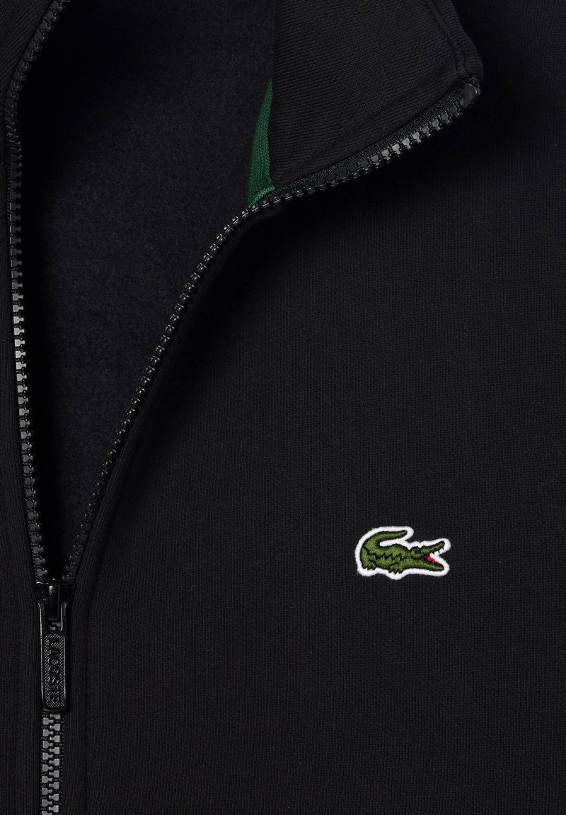 Schwarze Zip-Jacke mit einem grünen Krokodil-Logo, das auf der Brust bestickt ist, und einem teilweise geöffneten Kragen.