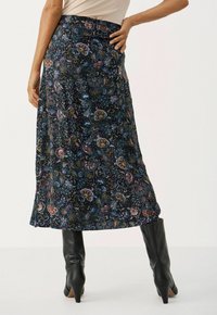 Donna che indossa una gonna midi nera a fiori abbinata a stivali con tacco neri, in piedi con una mano sulla vita.