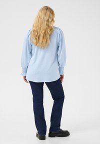 Blouse longue à manches bleues claires avec des manches froncées et une coupe ample, associée à un jean bleu foncé et des chaussures noires. Texture de tissu lisse.