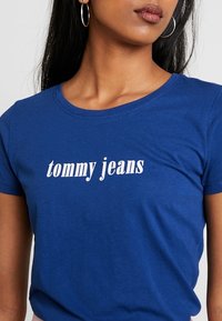 Blå bomullströja med korta ärmar, med texten "tommy jeans" i fet vit skrift över bröstet.