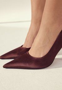 Bordo spalvos satino aukštakulniai bateliai su smailiu noseliu ir plačia iškirpte aplink pėdą. Lygi tekstūra ir elegantiškas dizainas pabrėžia rafinuotumą.