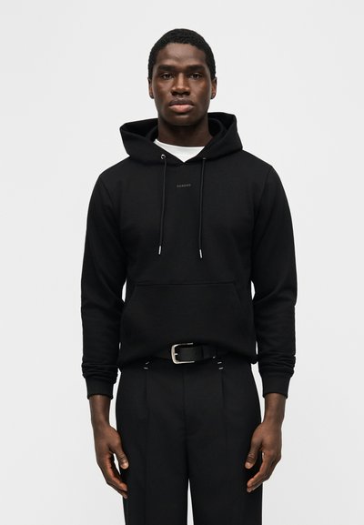 Νέος άνδρας που στέκεται με το πρόσωπο προς τα εμπρός, φορώντας μαύρο φούτερ με κουκούλα και μπροστινή τσέπη, και μαύρο παντελόνι με ζώνη, πάνω σε λευκό φόντο.