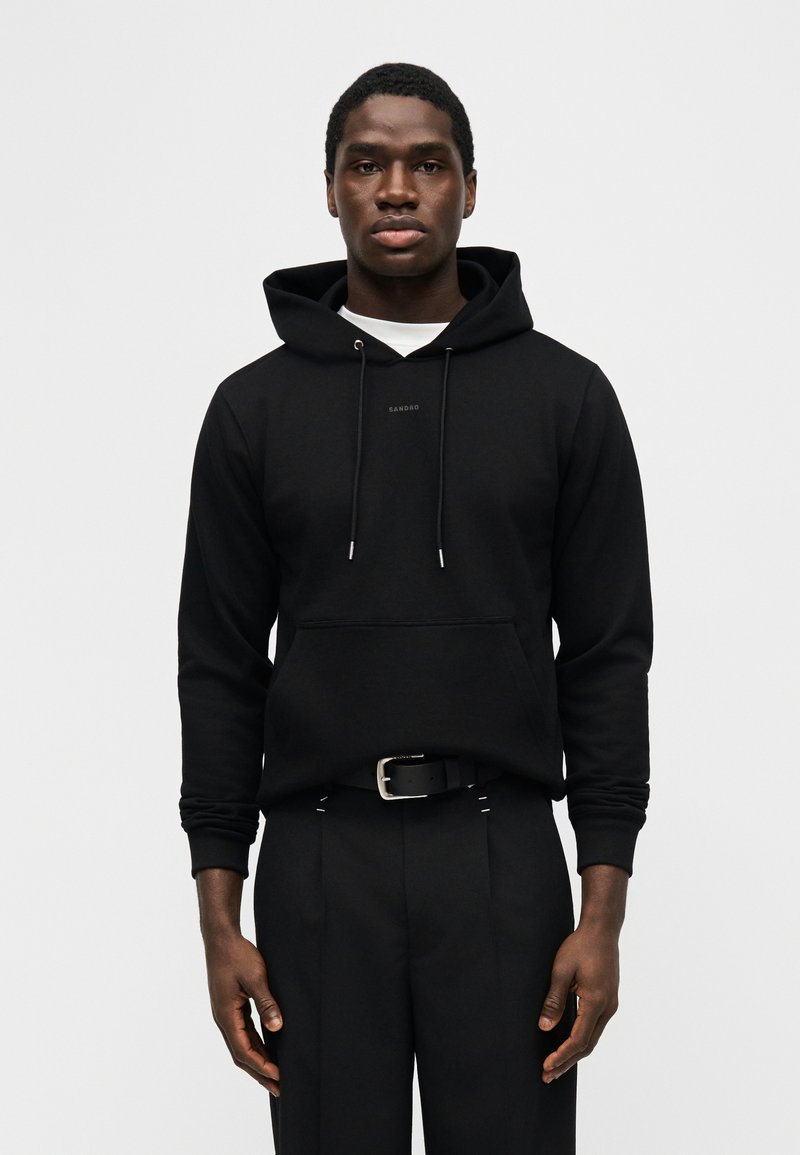 Junger Mann steht frontal, trägt einen schwarzen Kapuzenpullover mit Fronttasche und schwarze Hose mit Gürtel vor weißem Hintergrund.