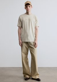 Bézs pamutpóló dombornyomott "BOSS" logóval, laza bézs nadrággal és hozzáillő sapkával, sima textúrával és kényelmes szabással.