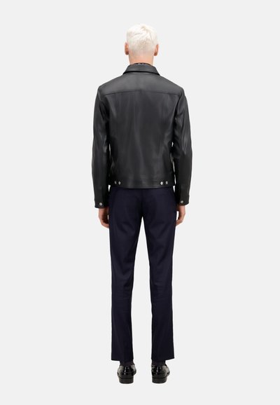 Giacca di pelle nera con colletto classico e bottoni a pressione, vista da dietro. Indossata sopra pantaloni blu navy e scarpe nere lucide.