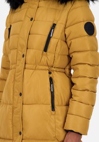 Mosterdgele pufferjack met een getailleerde taille, zwarte ritsen en een nepbont rand op de capuchon. Heeft een gequiltde textuur en een logo patch.