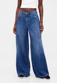 Modré džínové kalhoty s širokými nohavicemi a vysokým pasem, vybavené předními kapsami a klasickým zapínáním na knoflíky. Materiál má hladkou, mírně vybledlou texturu.