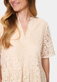 Lace dress in pale beige with floral pattern, short sleeves, and a v-neckline, featuring a tiered design and textured fabric.