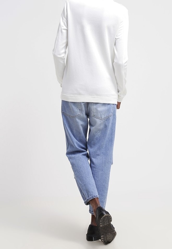 Vit långärmad topp med en avslappnad passform, tillsammans med ljusblå denimjeans med rak skärning och bakfickor. Svarta skor.