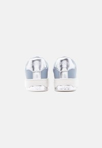 Ζευγάρι γκρι και ασημί αθλητικά παπούτσια, εμφανιζόμενα από την πίσω πλευρά, με λευκές σόλες και μεταλλικό λογότυπο "DKNY" στα τακούνια και στις μέσες σόλες.