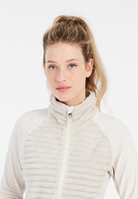 Fleece bunda svetlého béžového odtieňa s vysokým golierom, so vertikálnym rebrovaným vzorom na prednej strane a so zipsom. Mäkka a hladká látka.