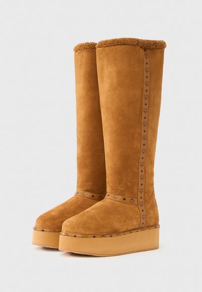 Bottes hautes jusqu'au genou en daim fauve avec un revers en fourrure douce, semelle plate à plateforme et branding décoratif sur le côté. Texture lisse avec une construction robuste.