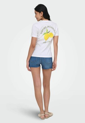 Kobieta w białej koszulce z nadrukiem cytryny i tekstem, szorty dżinsowe, stojąca tyłem na jednolitym tle.