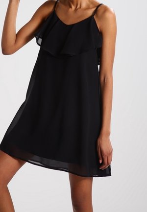 Femme portant une robe noire sans manches avec de fines bretelles et un col froncé sur un fond clair uni.