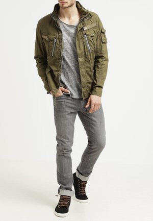 Chaqueta verde oliva con bolsillos con cremallera, camiseta gris, jeans gris claro y zapatillas negras con cordones marrones. Diseño de atuendo casual.