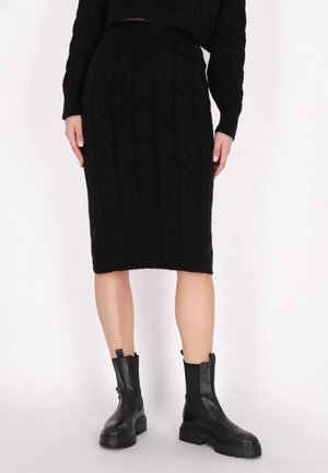 Jupe noire en maille torsadée longueur genou associée à des bottines noires épaisses sur fond blanc.