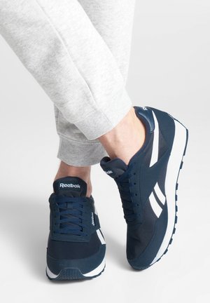 Persona che indossa scarpe da ginnastica Reebok blu navy e bianche con pantaloni della tuta grigi su uno sfondo chiaro uniforme.