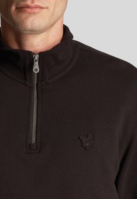 Schwarzer Quarter-Zip-Pullover aus weichem, strukturiertem Material mit einem gestickten Logo auf der linken Brust und einem silbernen Reißverschluss.