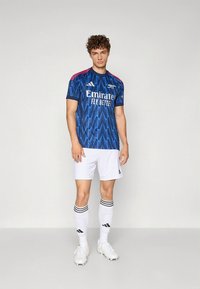 Camiseta de fútbol azul con patrón de zigzag, detalles rojos en los hombros, combinada con pantalones cortos blancos y calcetas a rayas. Corte y diseño atlético.