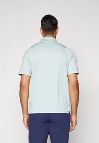 Man seen from behind wearing a light blue short-sleeve polo shirt and dark blue pants against a plain light background.