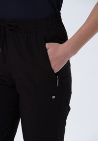 Pantalons de sport noirs avec une taille élastique, un cordon de serrage, des poches latérales zippées et un petit logo sur le bas de la jambe.