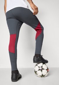 Pantalons de sport gris avec des accents rouges, design ajusté et tissu texturé, associés à des chaussures de football noires sur un ballon de football.