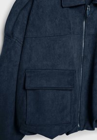 Tmavě modrá semišová bunda s předním zipem, velkou našitou kapsou a strukturovanými rameny, s jemnou texturou a minimalistickým designem.