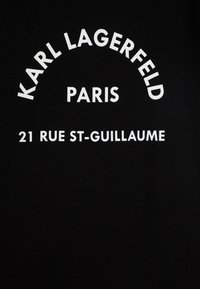 Juodas medvilnės marškinis, kuriame ryškus baltas tekstas: "KARL LAGERFELD PARIS" išlenktoje formoje ir "21 RUE ST-GUILLAUME" apačioje, paprastas ir modernus dizainas.