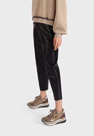 Baskets dorées métalliques avec des accents texturés, semelle blanche et lacets foncés, associées à un pantalon noir en simili cuir ajusté et un haut beige.