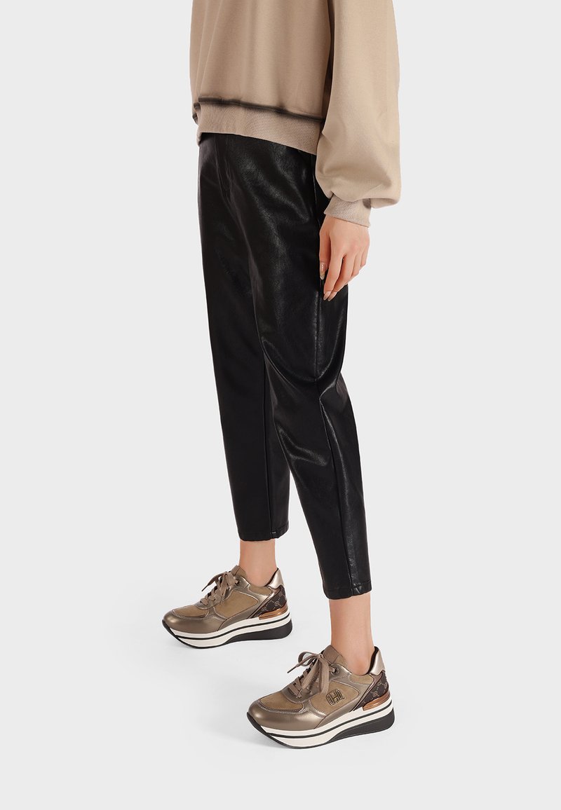 Sneakers dorati metallici con dettagli testurizzati, suola bianca e lacci scuri, abbinati a pantaloni tapered in ecopelle nera e una maglia beige.