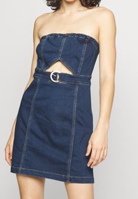Šaty bez ramínek z denimové látky v tmavě modré barvě, s výřezovým designem a kulatou stříbrnou sponou v oblasti pasu. Hladká textura, přiléhavý střih.