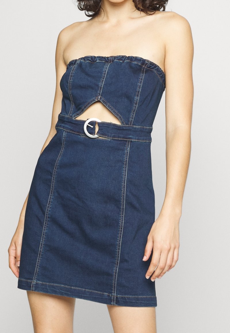 Šaty bez ramínek z denimové látky v tmavě modré barvě, s výřezovým designem a kulatou stříbrnou sponou v oblasti pasu. Hladká textura, přiléhavý střih.