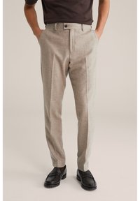 Bărbat purtând pantaloni bej croiți cu pliuri frontale, mocasini negri și o cămașă maro închis, stând cu mâinile în buzunare.